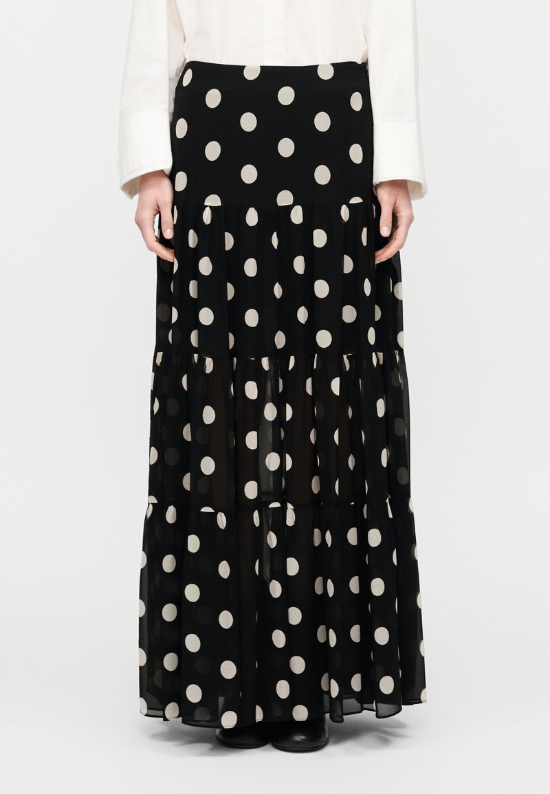 Μακριά φούστα από μαύρο διαφανές ύφασμα με μεγάλ α λευκά πουά, σχεδιασμός με στρώσεις και ελαστική ζώνη μέσης, συνδυασμένη με μαύρες μπότες.