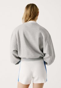 Felpa grigia crop con maniche oversize e polsini a coste, abbinata a pantaloni shorts bianchi con strisce laterali blu.