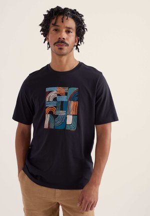 Mann mit lockigem Haar, der ein schwarzes T-Shirt mit abstrakten, bunten, geometrischen Mustern und beige Hosen vor einem einfarbigen Hintergrund trägt.