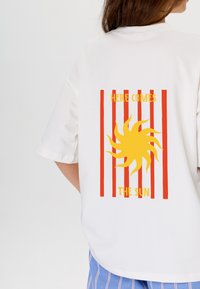 Witte T-shirt met korte mouwen, voorzien van een gele zonafbeelding met rode verticale strepen en de tekst "HIER KOMT DE ZON" eronder.