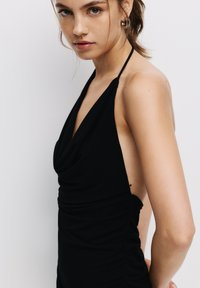 Robe noire sans manches avec un décolleté drapé et des fronces sur les côtés. Le tissu paraît lisse avec une texture douce et une coupe modeste sur les côtés.