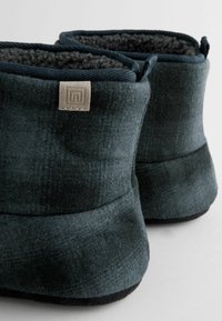 Încălțăminte de culoare verde închis, cu interior moale și pufos și exterior texturat. Dispune de o formă rotundă și de o etichetă mică cu logo pe spate.