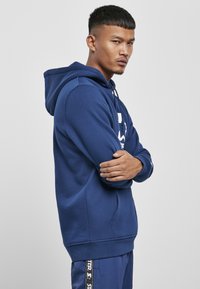 Kék kapucnis pulóver, kényelmes textúrával, elülső zsebbel és merész fehér logónyomattal. A modell karba font kézzel pózol.