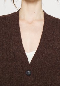 Hnědý pletený kardigan s výstřihem do V, vybavený jedním černým knoflíkem vpředu. Textura vypadá měkce a chlupatě.
