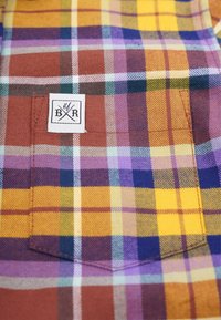 Tissu à carreaux avec des motifs bruns, jaunes et violets. Comprend une étiquette cousue avec des initiales sur la poche de poitrine. Matériau texturé.