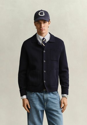 Homme portant une casquette bleu marine avec un grand G, un cardigan bleu marine, une cravate rayée, une chemise à boutons, un jean et une montre, debout devant un fond uni.