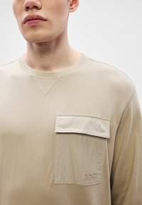 Béžová košeľa s dlhými rukávmi a textúrovanou látkovou náprsnou kapsou na ľavej strane. Má okrúhly výstrih a jemné rebrovanie.