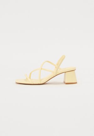 Beige sandal med tynde stropper og lav blokhæl, vist fra siden på en ensfarvet lys baggrund.