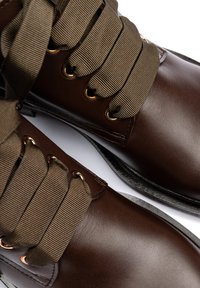 Brown leather boots feature wide, olive green ribbon laces, metal eyelets, and a smooth, glossy finish. Sole is slightly raised.
