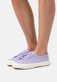 Sneakers in tela lavanda con suole e lacci bianchi, caratterizzati da un upper testurizzato e una patch con il logo sul lato.