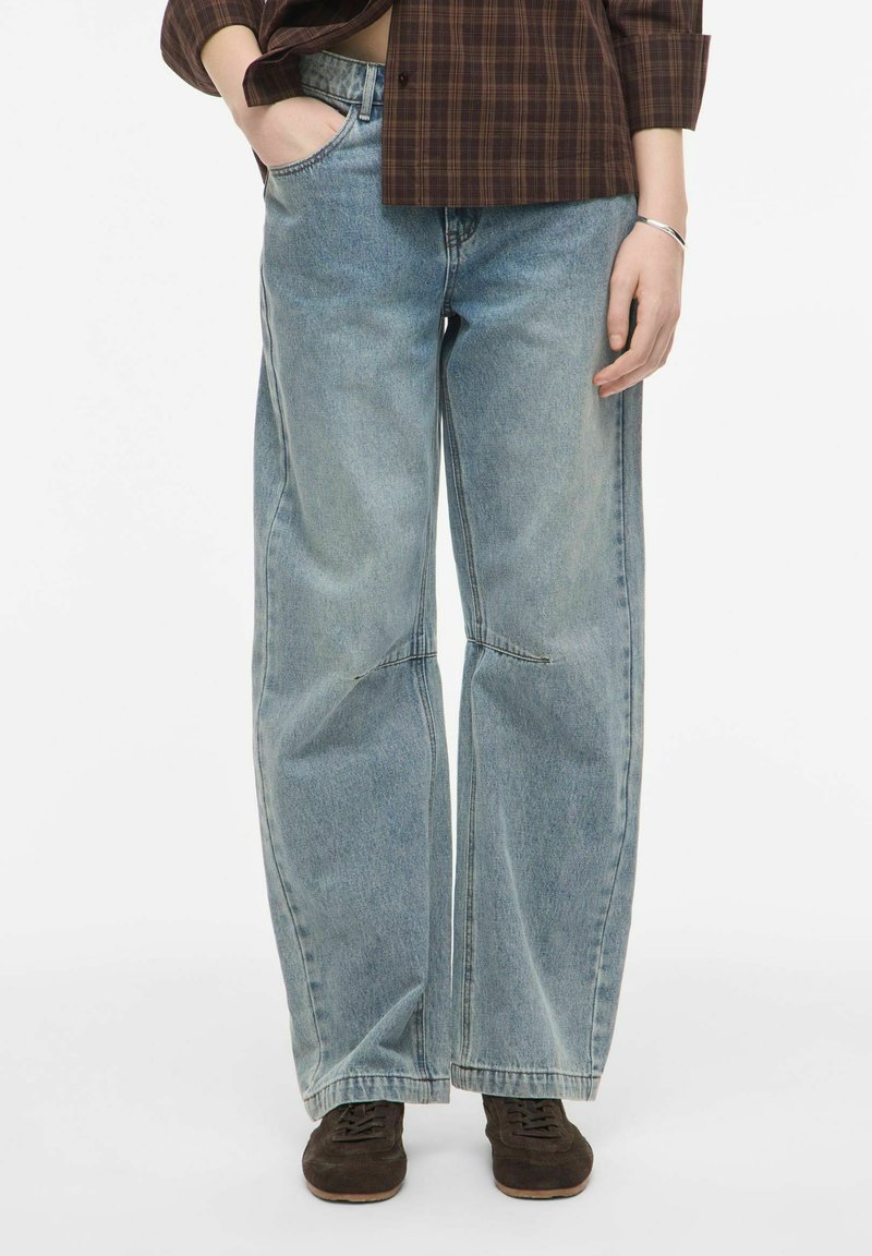 Personne portant un jean large bleu clair, une chemise à carreaux marron et des chaussures marron foncé, debout avec une main dans la poche.