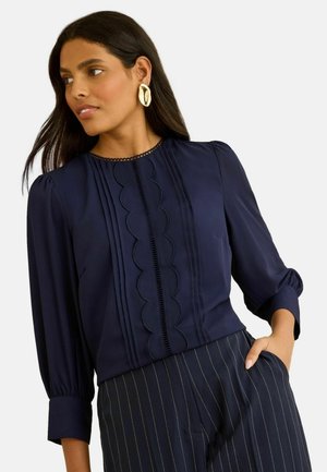 Žena nosící námořnicky modrou halenku s vroubkovanými a plisovanými detaily vpředu, nafouklými manžetami a tmavě modré kalhoty s tenkými pruhy, s rukou v kapse.