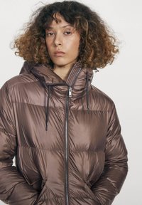 Kvinde med krøllet hår iført en skinnende brun dunjakke med høj krave og sorte snore, stående mod en ensartet lys baggrund.