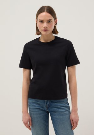 Mujer joven con cabello corto y castaño, vistiendo una camiseta negra sencilla de manga corta y jeans azules, de pie contra un fondo claro.