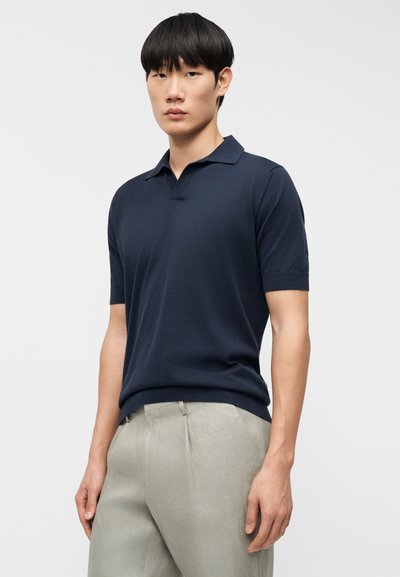 Μπλε ναυτικό κοντομάνικο πόλο πουκάμισο με γιακά και ίσια γραμμή. Μαλακό, υφασμάτινο με υφή; σετ με ανοιχτόχρωμο, πλισέ παντελόνι.