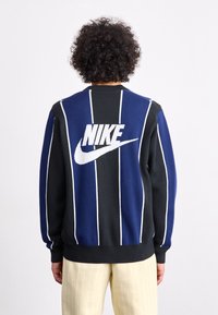 Nike-neulottu paita, jossa on pystysuorat raidat laivastonsinisinä ja mustina, selässä näkyy suuri valkoinen logo, sekä resorit hihansuissa ja helmassa.