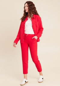 Rode overhemd met knopen en twee voorzakken, gecombineerd met bijpassende slim-fit broeken. Witte shirt eronder en witte sneakers met zwarte accenten.