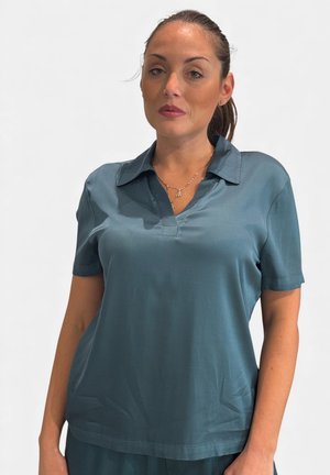 Donna con capelli raccolti che indossa una camicia verde acqua a maniche corte con colletto e una collana d'oro, in piedi davanti a uno sfondo chiaro e uniforme.