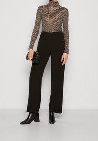 Svarta högmidjade byxor i kombination med en mönstrad, figursydd polotröja i beige och brunt. Modellen håller en liten svart handväska.