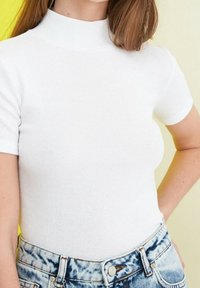 Žena v upnutém bílém topu s krátkým rukávem a vysokými modrými džínami s efektem acid wash, stojí s jednou rukou na boku.
