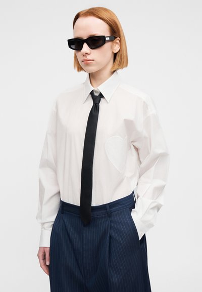 Λευκό πουκάμισο με γιακά και καρδιά σαν επιδιορθωτική λεπτομέρεια, συνδυασμένο με μαύρη γραβάτα και ναυτικοδίχρωμο παντελόνι με λεπτές ρίγες. Μαύρα γυαλιά ηλίου ολοκληρώνουν την εμφάνιση.