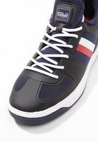 Tummansininen lenkkari, jossa on kuminen varvaskuppi, verkkomateriaali päällisessä ja valkoiset nauhat. Sivulla on valkoiset ja punaiset raidat sekä logo kielekkeessä.