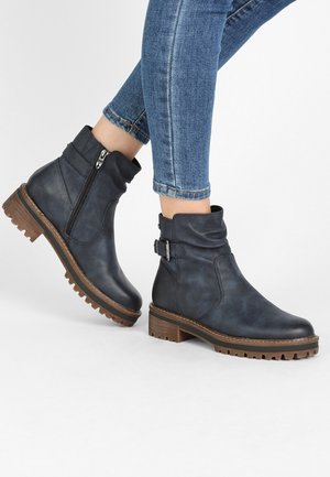 Jambes en jean bleu portant des bottines en cuir noir avec boucles, fermetures éclair latérales et semelles épaisses marron à crampons.