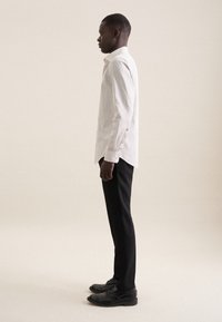 Chemise blanche à motifs à manches longues avec un col boutonné, assortie à un pantalon noir sur mesure et des chaussures noires, vue de côté.