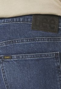 Jeans de mezclilla azul oscuro con una etiqueta de cuero negro texturizado que muestra "Lee" en la cintura y una pequeña etiqueta dorada en el bolsillo trasero.