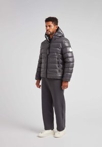 Chaqueta de plumas gris con textura acolchada, dos bolsillos frontales, capucha con detalles de logo y cremallera negra. Combinada con pantalones grises y zapatillas blancas.
