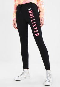 Osoba ubrana w czarne legginsy Hollister z różowym logo na lewej nodze, różowo-żółty sweter w stylu tie-dye oraz białe sneakersy za kostkę.