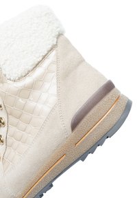 Botte d'hiver beige avec une doublure en fausse fourrure, panneaux latéraux texturés, œillets dorés et une semelle en caoutchouc brun et gris avec un motif de crampons.