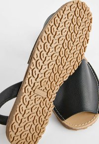 Sort lædersandal med en tekstureret tan gummisål, der har et mønstret greb. Syninger langs kanten fremhæver designet.