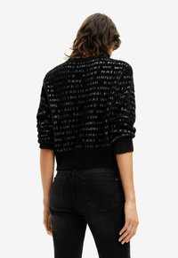 Czarny, puszysty sweter z teksturowanym materiałem, na którym znajdują się subtelne wzory tekstowe. Dzwonki i krawędź z prążkowanym wykończeniem; zestawiony z ciemnymi dżinsami.