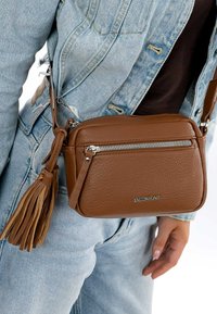 Pruun nahk crossbody kott, millel on tekstureeritud pind, hõbedane tõmblukk ja tuti detail, sinise tekstiili rõivaste taustal.