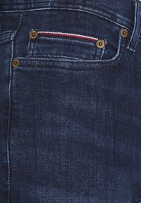 Mörk denimjeans med en liten framficka med dekorativ sömnad i röd och vit, guldiga detaljer vid fickans kanter.