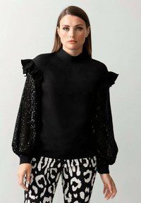 Schwarzes Oberteil mit Rüschenschultern und gepailletten Ballonärmeln, kombiniert mit Hosen im Schwarz-Weiß-Muster. Insgesamt glatte Textur.
