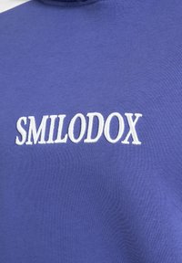 Blue sweatshirt featuring white embroidered text "SMILODOX" on the chest. Material appears soft with a smooth texture.