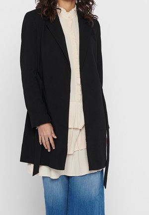 Kvinde iført en sort blazer med åben front over en cremefarvet bluse med flæser og blå denim jeans, stående mod en ensfarvet lys baggrund.