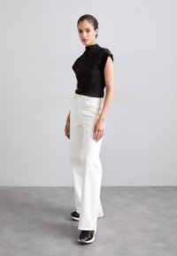 Haut noir sans manches à col montant ; jean blanc à jambes larges ; baskets noires avec semelle crème ; le modèle pose devant un fond neutre.