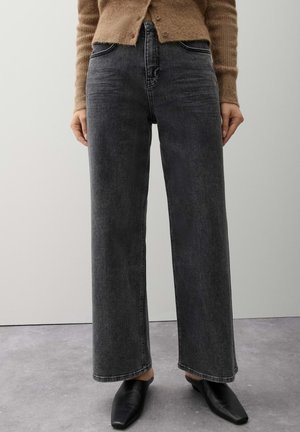 Persoon die donkergrijze, high-waisted, wijde jeans draagt, gecombineerd met zwarte puntige schoenen en een bruine gebuttonede trui.