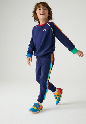 Felpa blu navy con dettagli arcobaleno, accenti a righe colorate sulle maniche, pantaloni coordinati. Sneakers dai colori vivaci con doppie cinghie.