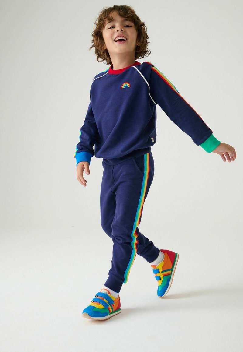Felpa blu navy con dettagli arcobaleno, accenti a righe colorate sulle maniche, pantaloni coordinati. Sneakers dai colori vivaci con doppie cinghie.