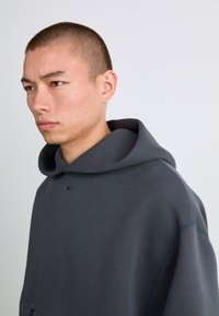 Mørkegrå hættetrøje med en stor, struktureret hætte, blød tekstur og lille Nike-logo på brystet. Har en løs pasform og minimale syninger.
