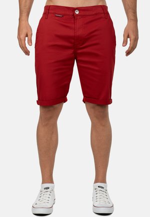 Mann trägt hochgekrempelte rote Shorts und weiße Sneaker mit niedrigem Schaft und steht vor einem einfarbigen Hintergrund.