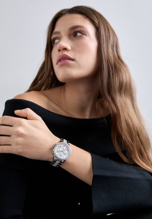 Jeune femme aux longs cheveux bruns, portant un haut noir épaules dénudées et une montre argentée au poignet, regardant pensivement vers sa gauche sur un fond uni.