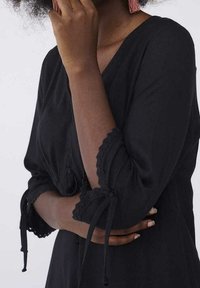 Svart klänning med långa ärmar som har spetskant och knytningar. Slät tyg. Nära bild av arm och hand som vilar över kroppen.