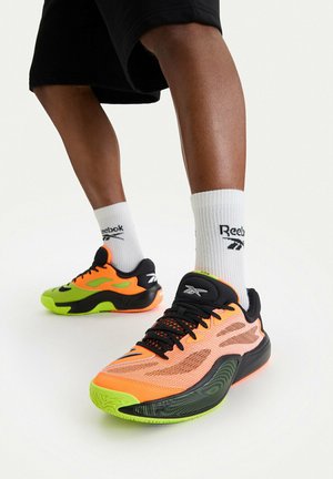 Persona lleva pantalones cortos negros, calcetines Reebok blancos hasta la pantorrilla y zapatillas Reebok de neón naranja y verde, de pie sobre un suelo blanco.