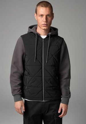 Gilet matelassé noir avec fermeture par zip et capuche, avec des manches grises texturées, des poignets élastiques et des poches latérales. Conçu pour le confort et la chaleur.