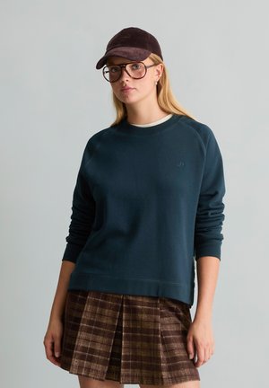 Jeune femme portant une casquette sombre, de grandes lunettes, un sweat-shirt bleu marine et une jupe plissée à carreaux marron, debout devant un fond uni.
